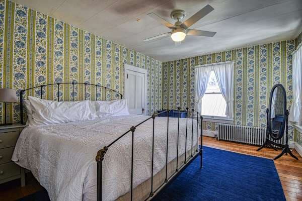【Queen Bedroom 2】 Queen iron bed, green-and-white striped wallpaper, full-length mirror — blue rug, radiator heat.