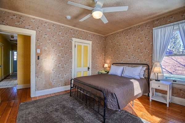 【Queen Bedroom 1】 Queen iron bed, pink-floral wallpaper, yellow Victorian door — original 1880s detail, remodeled comfort.